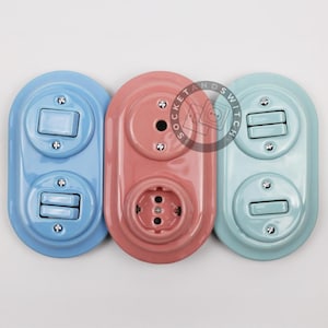 May include: Three oval-shaped wall plates with electrical components. The left plate is light blue with two toggle switches. The center plate is dusty pink with a circular outlet and a blank cover. The right plate is mint green with two switches.