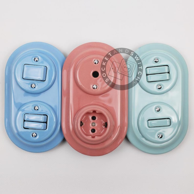 May include: Three oval-shaped wall plates with electrical components. The left plate is light blue with two toggle switches. The center plate is dusty pink with a circular outlet and a blank cover. The right plate is mint green with two switches.