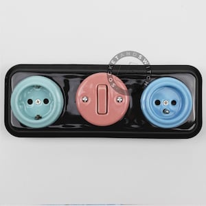 May include: A rectangular black wall plate featuring a light blue electrical outlet on the left, a round pink light switch in the center, and a medium blue electrical outlet on the right. The components are mounted on a glossy black base.