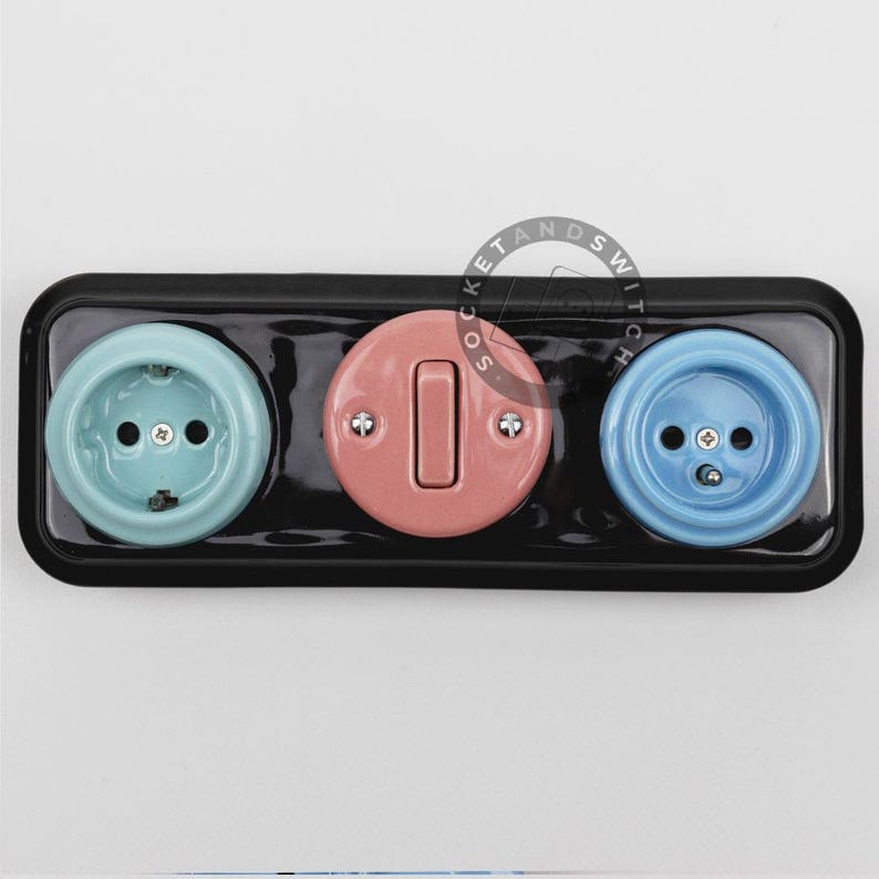 May include: A rectangular black wall plate featuring a light blue electrical outlet on the left, a round pink light switch in the center, and a medium blue electrical outlet on the right. The components are mounted on a glossy black base.