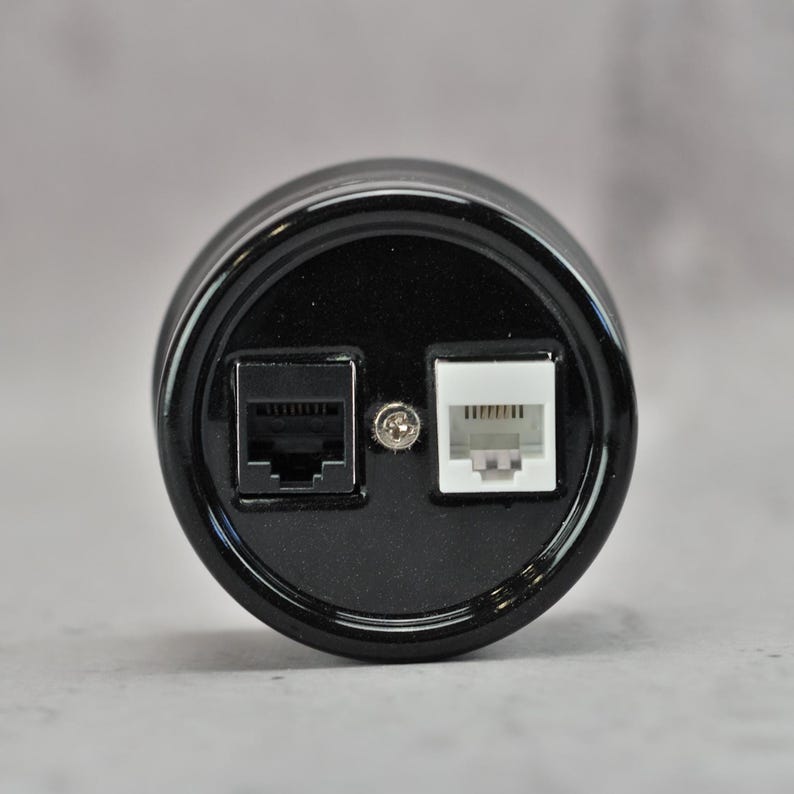 May include: A circular black electronic component featuring a glossy finish with one black RJ45 ethernet port and one white RJ11 telephone port positioned side by side, separated by a small silver metal screw in the center of the faceplate.