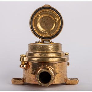 May include: A brass-coloured industrial component with an open, circular lid. The component has a cylindrical body with a protruding nozzle and two rectangular base extensions. The lid is hinged and has a black rubber seal.