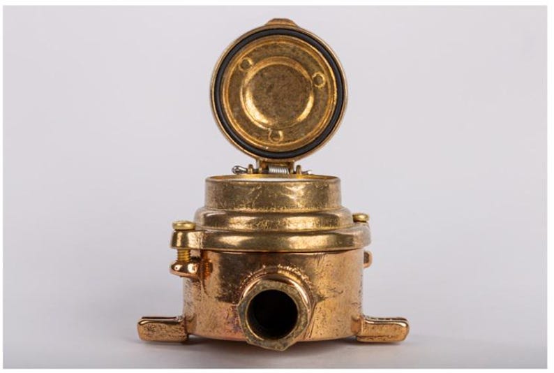 May include: A brass-coloured industrial component with an open, circular lid. The component has a cylindrical body with a protruding nozzle and two rectangular base extensions. The lid is hinged and has a black rubber seal.