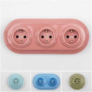 以下が含まれることがあります： ピンク色の電気コンセントで、3つの丸いソケットがあります。 コンセントは丸みを帯びた長方形の形をしており、マットな仕上がりです。 ソケットには真鍮色のピンと黒い穴があります。 青と緑のオプションも表示されています。