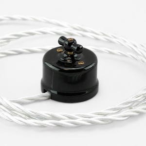 May include: A round black light switch with a central rotating toggle handle and two small brass screws on the top surface. The switch is connected to a long, white, twisted fabric-covered electrical cord that coils around the base.