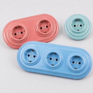 Può includere: Una collezione di tre prese elettriche a muro in ceramica con finitura lucida. Una presa doppia è rosa corallo, una presa singola è turchese chiaro e una presa tripla è azzurro cielo, ognuna con aperture circolari standard.