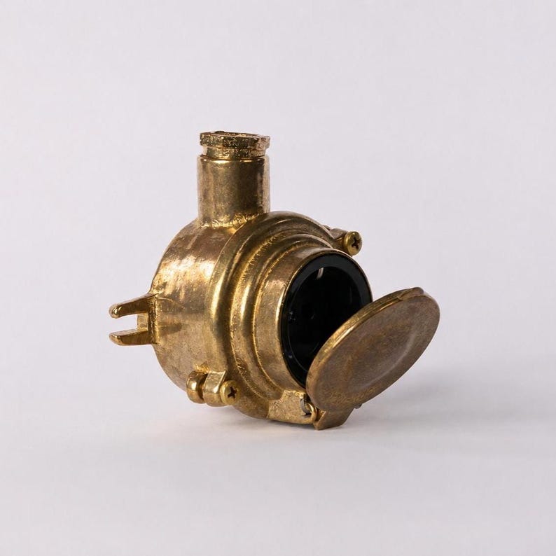May include: A brass-coloured, industrial-style object with a circular body and a hinged cover. The object has a cylindrical top and a dark interior visible through the open cover. The design suggests a vintage or nautical theme.