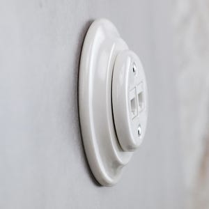 May include: A white ceramic wall-mounted dual data port outlet featuring a stepped, circular design. The glossy finish highlights the two rectangular ports positioned vertically, secured by two small silver-coloured metal screws on the faceplate.