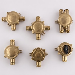 May include: Six brass electrical junction boxes are displayed. The boxes have a circular design with multiple entry points for wiring. Some have screw-on covers, and one has a black switch.