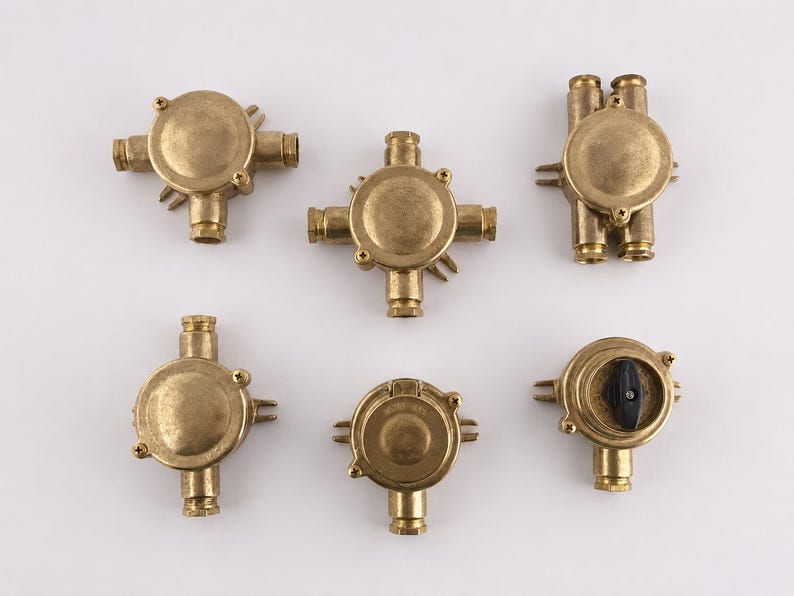 May include: Six brass electrical junction boxes are displayed. The boxes have a circular design with multiple entry points for wiring. Some have screw-on covers, and one has a black switch.