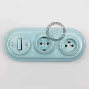 May include: A light blue, oval-shaped ceramic wall plate featuring one toggle light switch and two electrical outlets. The surface has a glossy finish with visible screws securing the components. A circular logo with the text SOCKET AND SWITCH is visible.