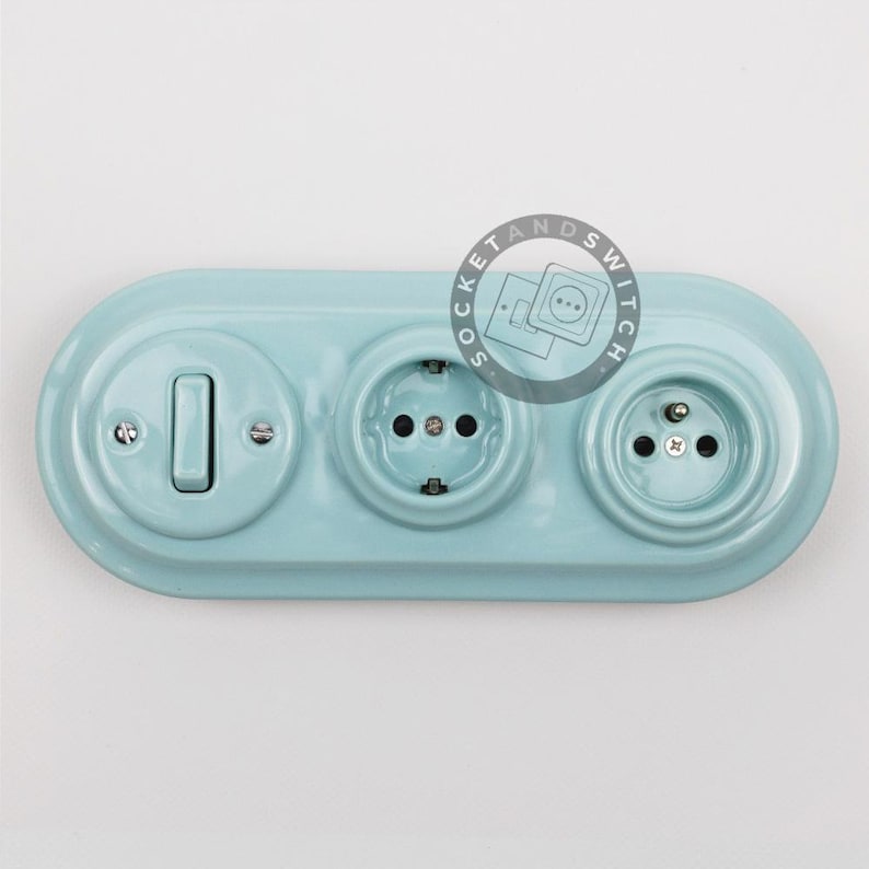May include: A light blue, oval-shaped ceramic wall plate featuring one toggle light switch and two electrical outlets. The surface has a glossy finish with visible screws securing the components. A circular logo with the text SOCKET AND SWITCH is visible.
