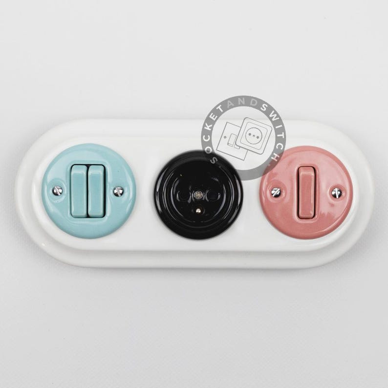 May include: A white oval wall plate featuring three circular inserts: a light blue double toggle switch on the left, a black electrical outlet in the center, and a pink single toggle switch on the right. The text SOCKET AND SWITCH is visible.