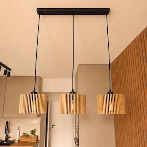 Puede incluir: Una lámpara colgante de tres luces con un soporte rectangular negro en el techo y tres cables negros que sostienen pantallas cilíndricas. Cada pantalla está construida con listones de madera clara en posición vertical.