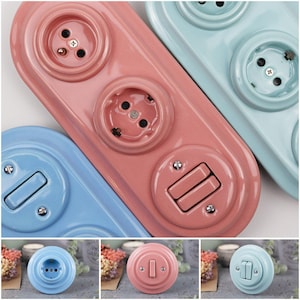 Könnte beinhalten: Eine Sammlung von elektrischen Steckdosen und Lichtschaltern aus Keramik in Pastellfarben. Die Steckdosen sind rosa, blau und hellblau, mit einem Vintage-Look. Die Schalter und Steckdosen haben eine glänzende Oberfläche und sind auf rechteckigen oder runden Platten montiert.