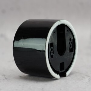 May include: A cylindrical black ceramic wall mount base with a glossy finish and a white rim. The interior features a black plastic insert with a U-shaped cutout and multiple small circular screw holes for mounting purposes.