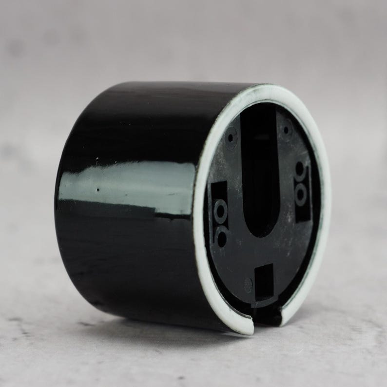 May include: A cylindrical black ceramic wall mount base with a glossy finish and a white rim. The interior features a black plastic insert with a U-shaped cutout and multiple small circular screw holes for mounting purposes.