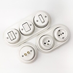 May include: A collection of white ceramic electrical components including a triple-switch wall plate, a double-outlet wall plate, and a single circular base with two brass contact pins. Each piece features a glossy finish and exposed metal screws.