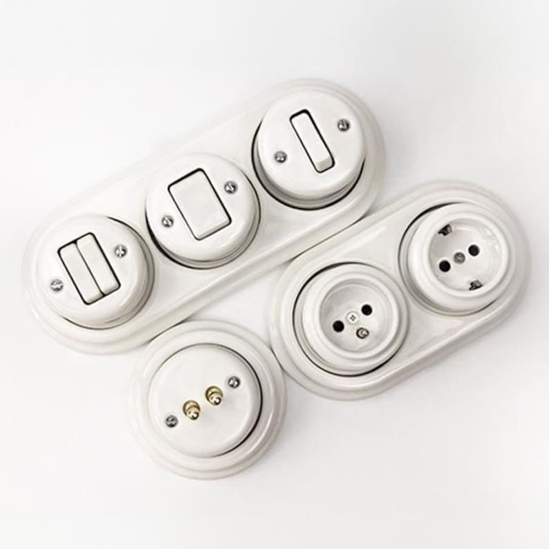 May include: A collection of white ceramic electrical components including a triple-switch wall plate, a double-outlet wall plate, and a single circular base with two brass contact pins. Each piece features a glossy finish and exposed metal screws.