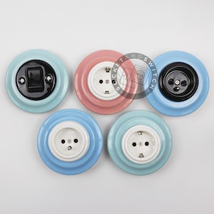 May include: Five circular wall-mounted electrical fixtures with glossy ceramic bases in pastel blue, pink, and light blue. The fixtures include a black toggle switch, a black power outlet, and three white power outlets, each featuring visible metal screws.