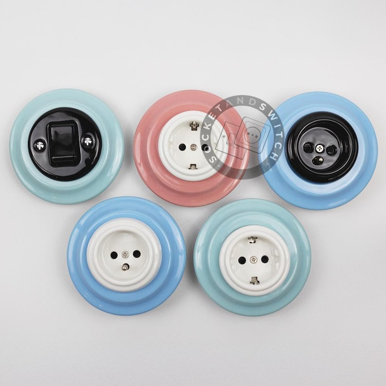 May include: Five circular wall-mounted electrical fixtures with glossy ceramic bases in pastel blue, pink, and light blue. The fixtures include a black toggle switch, a black power outlet, and three white power outlets, each featuring visible metal screws.