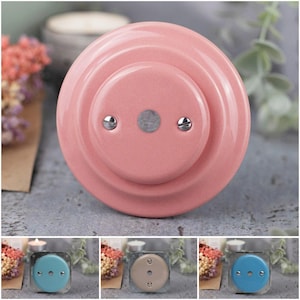 May include: A pink ceramic light switch cover with two silver screws and a central metal disc. The cover has a circular design with a raised center. Additional covers in blue and beige are shown below.