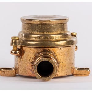 May include: A vintage brass fitting with a cylindrical body, a circular top, and a prominent central opening. The fitting has a weathered appearance, with visible screw heads and mounting tabs. The overall design suggests a functional, industrial aesthetic.