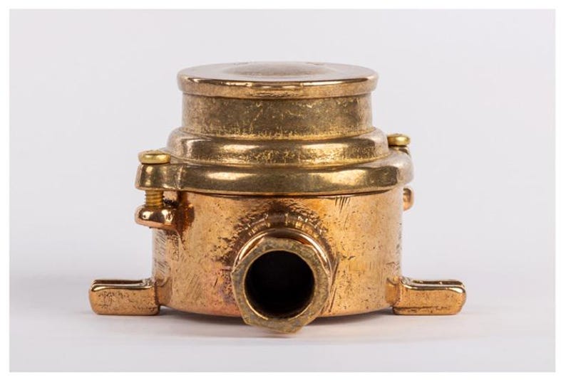 May include: A vintage brass fitting with a cylindrical body, a circular top, and a prominent central opening. The fitting has a weathered appearance, with visible screw heads and mounting tabs. The overall design suggests a functional, industrial aesthetic.