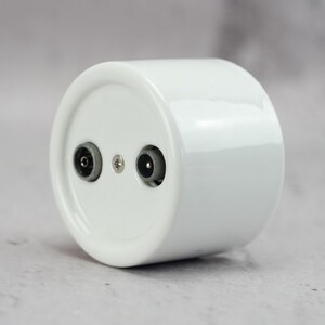May include: A cylindrical white ceramic electrical wall socket component featuring two circular ports and a central metal screw. The smooth, glossy finish of the white housing contrasts with the dark grey interior of the two electrical connection points.
