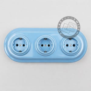 May include: A light blue triple electrical wall outlet with a glossy finish and a rounded rectangular shape. The unit features three circular sockets arranged horizontally, each with two pin holes and a central screw. A circular logo reads SOCKETANDSWITCH.