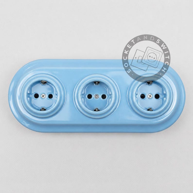 May include: A light blue triple electrical wall outlet with a glossy finish and a rounded rectangular shape. The unit features three circular sockets arranged horizontally, each with two pin holes and a central screw. A circular logo reads SOCKETANDSWITCH.