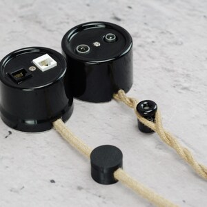 May include: Two cylindrical black wall sockets with glossy finishes. One unit features a white and a black modular port, while the other has two circular coaxial connectors. Both are connected to a twisted beige fabric cord with black accents.