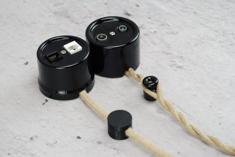May include: Two cylindrical black wall sockets with glossy finishes. One unit features a white and a black modular port, while the other has two circular coaxial connectors. Both are connected to a twisted beige fabric cord with black accents.
