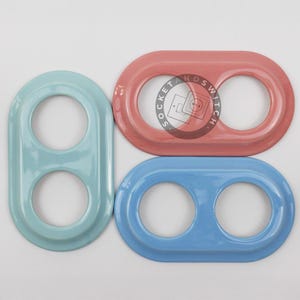 May include: Three oval-shaped double light switch plates with a glossy finish. One plate is light blue, one is a muted coral pink, and one is a medium blue. Each plate features two circular cutouts for switches and a circular logo with the text SOCKET AND SWITCH.