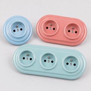 Può includere: Una collezione di tre prese elettriche a muro con finitura lucida. Una presa singola rotonda azzurra, una presa doppia ovale rosa corallo e una presa tripla ovale verde acqua chiaro, ciascuna dotata di un perno di messa a terra centrale.
