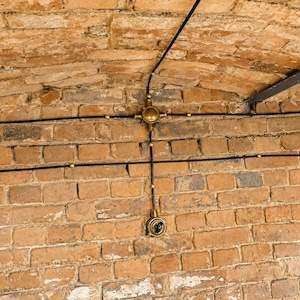 May include: A close-up of a brick wall with exposed electrical wiring. The black wiring runs horizontally and vertically, connected to brass fixtures. The bricks are a mix of orange and brown tones, creating a rustic aesthetic.