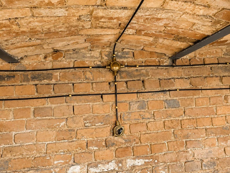 May include: A close-up of a brick wall with exposed electrical wiring. The black wiring runs horizontally and vertically, connected to brass fixtures. The bricks are a mix of orange and brown tones, creating a rustic aesthetic.