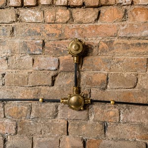 May include: An industrial electrical fitting mounted on a weathered brick wall. The fixture has brass-coloured components and black wiring, creating a vintage aesthetic. The brick wall has a mix of brown and tan tones.