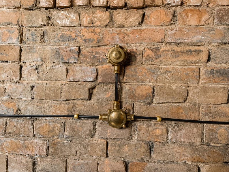 May include: An industrial electrical fitting mounted on a weathered brick wall. The fixture has brass-coloured components and black wiring, creating a vintage aesthetic. The brick wall has a mix of brown and tan tones.