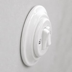 May include: A white ceramic light switch with a circular base plate and a single toggle lever. The switch features a glossy finish and is secured with a visible metal screw on the side of the central circular housing, showing a side profile view.