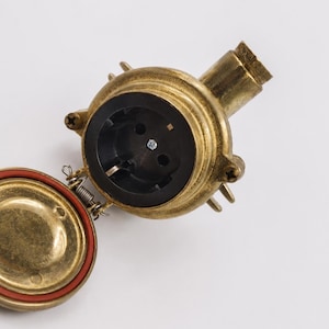May include: A brass electrical socket with a black interior and a hinged cover with a red seal. The socket has a cylindrical shape with a threaded end and is mounted on a white surface.