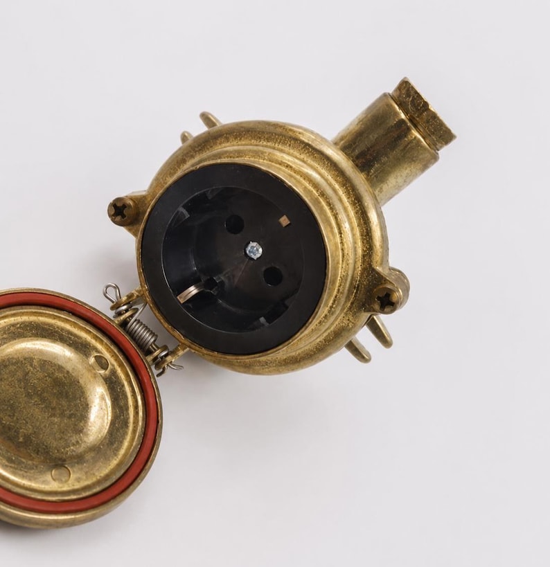 May include: A brass electrical socket with a black interior and a hinged cover with a red seal. The socket has a cylindrical shape with a threaded end and is mounted on a white surface.