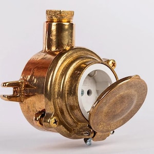 May include: A brass electrical socket with a hinged cover. The socket has a circular design and a white interior. The brass has a warm, aged appearance, and the socket is designed for durability. The socket is suitable for use in the UK.