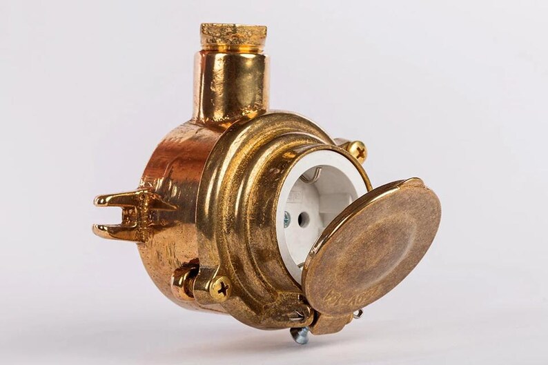 May include: A brass electrical socket with a hinged cover. The socket has a circular design and a white interior. The brass has a warm, aged appearance, and the socket is designed for durability. The socket is suitable for use in the UK.