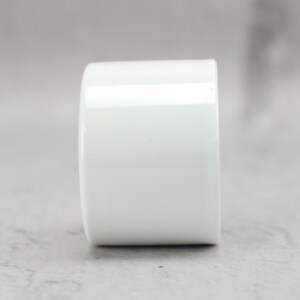 May include: A cylindrical white ceramic object with a smooth, glossy finish. The item features a simple, uniform shape with a flat top and bottom, presenting a clean, solid white surface that reflects light evenly across its curved exterior.