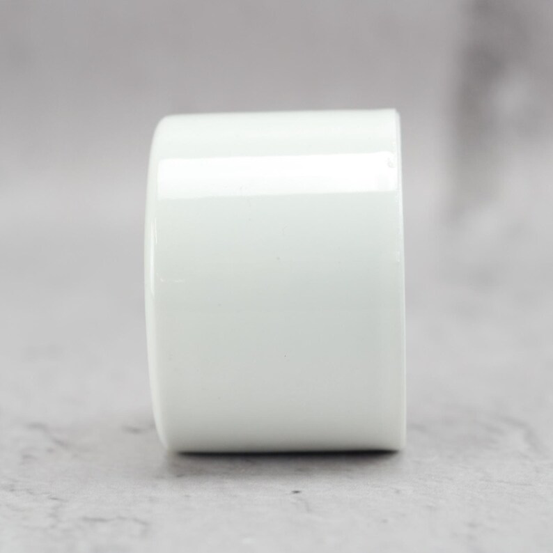 May include: A cylindrical white ceramic object with a smooth, glossy finish. The item features a simple, uniform shape with a flat top and bottom, presenting a clean, solid white surface that reflects light evenly across its curved exterior.