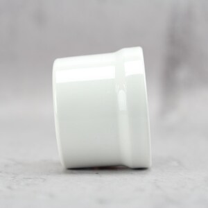May include: A small, cylindrical white ceramic ramekin with a smooth, glossy finish. The vessel features a slightly flared rim at the top and a straight-sided body, presenting a clean, minimalist design suitable for serving individual portions of food.