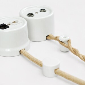 May include: Two white cylindrical wall-mounted electrical outlet units with ports for data and coaxial cables. Each unit features a side opening for a beige twisted fabric-covered electrical cord, accompanied by small white ceramic cord guides.