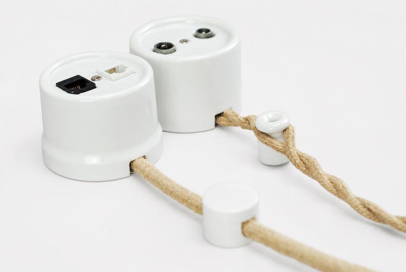 May include: Two white cylindrical wall-mounted electrical outlet units with ports for data and coaxial cables. Each unit features a side opening for a beige twisted fabric-covered electrical cord, accompanied by small white ceramic cord guides.