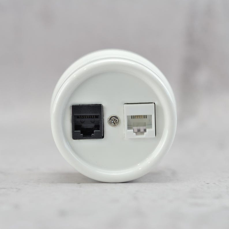 May include: A circular white wall plate featuring two modular ports. One port is black and the other is white, positioned side-by-side. A small silver metal screw is located in the center between the two ports, securing the faceplate in place.